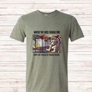 May include: Green t-shirt featuring a graphic from "Where the Wild Things Are." The illustration shows a creature sitting near a sailboat and palm trees. Text on the shirt reads "Where the Wild Things Are" and "Story and Pictures by Maurice Sendak."