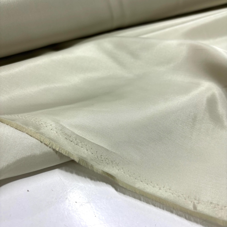 May include: A roll of off-white fabric with a smooth, shiny surface. The fabric is folded and has a visible seam along the edge. The material appears to be lightweight and suitable for clothing or decorative projects.