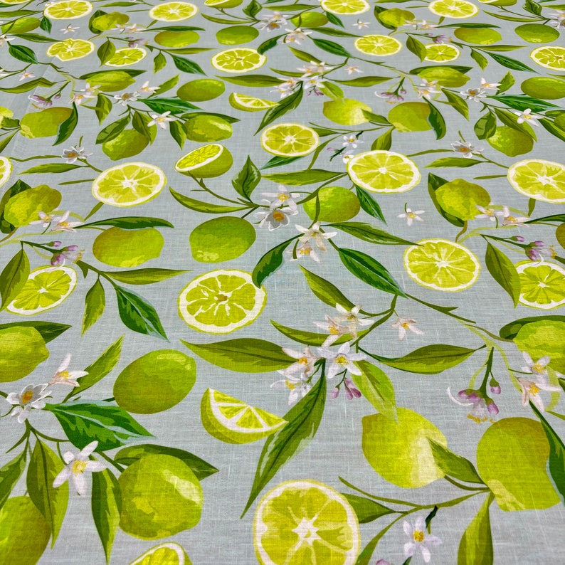 May include: A light blue fabric with a repeating pattern of green limes, lime slices, green leaves, and white flowers. The limes are whole, sliced, and in various orientations, creating a fresh, summery design. The fabric is likely cotton or linen.