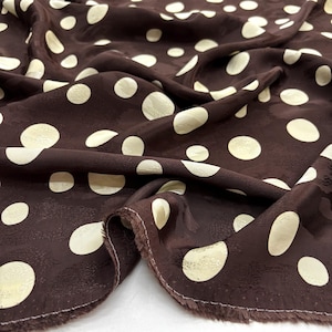 May include: Close-up of a brown fabric with a pattern of large, cream polka dots. The fabric is draped, showing folds and texture. The edges of the fabric are frayed.