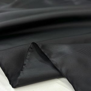 Puede incluir: Primer plano de tela negra con un ligero brillo. La tela está doblada, revelando su textura y caída. El color oscuro y la superficie lisa sugieren un textil lujoso, adecuado para ropa o decoración del hogar.