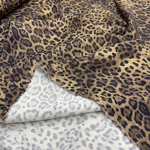 May include: A close-up shot of leopard print fabric. The fabric has a tan base with dark brown spots. The reverse side of the fabric is light grey with a less defined leopard pattern.