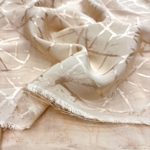 May include: Cream-coloured fabric with a metallic gold geometric pattern. The fabric is draped, showing the texture and sheen of the material. The edges are frayed, adding a delicate touch. Suitable for apparel or home decor.