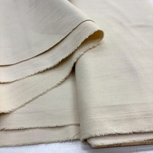 Op de afbeelding: Een lengte ongebleekte, off-white katoenen stof met een zichtbare geweven textuur en rauwe, gerafelde randen. Het materiaal is in lagen gevouwen en vertoont een consistente lichtcrème kleur over het gehele textieloppervlak en het weefsel.