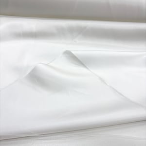 May include: Close-up of a roll of white satin fabric. The fabric is folded and draped, showing the smooth texture and sheen. The folds create shadows and highlights, emphasising the fabric's luxurious appearance.