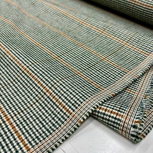Könnte beinhalten: Nahaufnahme eines gefalteten Stoffes mit einem grünen, weißen und braunen Karomuster. Der Stoff hat eine strukturierte Oberfläche mit diagonalen und vertikalen Streifen. Die Kanten des Stoffes sind sichtbar und zeigen das Gewebe und die Schichten des Materials. Dieser Stoff eignet sich für Kleidung und Heimdekoration.