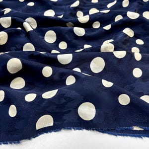 May include: Dark blue fabric featuring a pattern of large, cream-coloured polka dots. The material is textured and draped, with visible folds and shadows. The edges of the fabric are visible, indicating it's a cut piece.