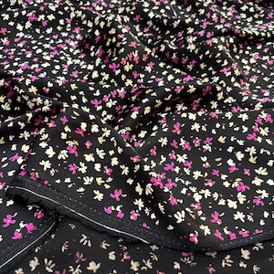 May include: Black fabric covered in a repeating floral design. The small flowers are in pink, white, and yellow. The fabric is folded, revealing the edge with a white border.