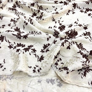 May include: Cream-coloured fabric featuring a repeating pattern of dark brown floral branches and leaves. The fabric is draped, showing folds and texture. The floral design covers the entire surface.