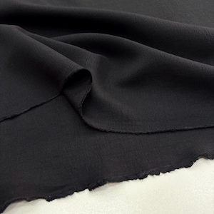 Op de afbeelding: Close-up van zwarte stof met een getextureerd oppervlak. De stof is gedrapeerd en toont plooien en een ruwe, onafgewerkte rand. Het materiaal lijkt zacht en licht, geschikt voor kleding of knutselprojecten.