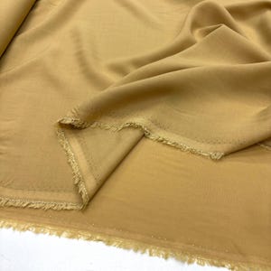Puede incluir: Primer plano de una tela color beige con una textura suave y fluida. La tela está doblada, revelando un borde deshilachado con pequeños agujeros espaciados uniformemente. El color es un tono cálido y neutro, adecuado para varios proyectos de costura.
