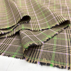 May include: Close-up of a folded fabric with a plaid pattern. The fabric features a tan base with black, green, and purple intersecting lines. The edges of the fabric are frayed. The fabric is likely used for clothing or home decor.