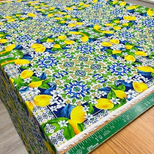 May include: White fabric with a repeating pattern of blue and green tiles, yellow lemons, and white flowers. The fabric has a white fringe along the edge.