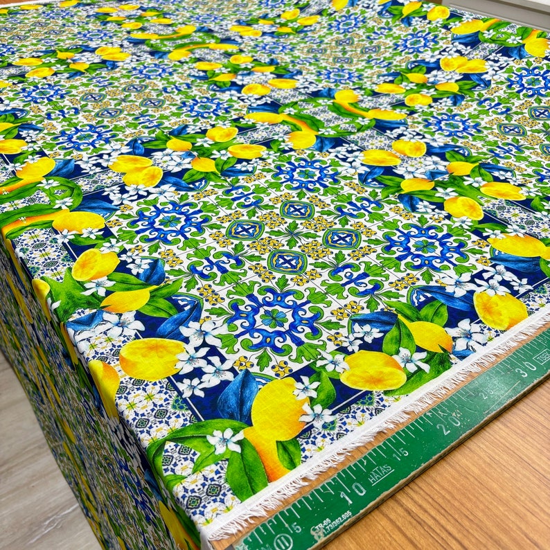 May include: White fabric with a repeating pattern of blue and green tiles, yellow lemons, and white flowers. The fabric has a white fringe along the edge.