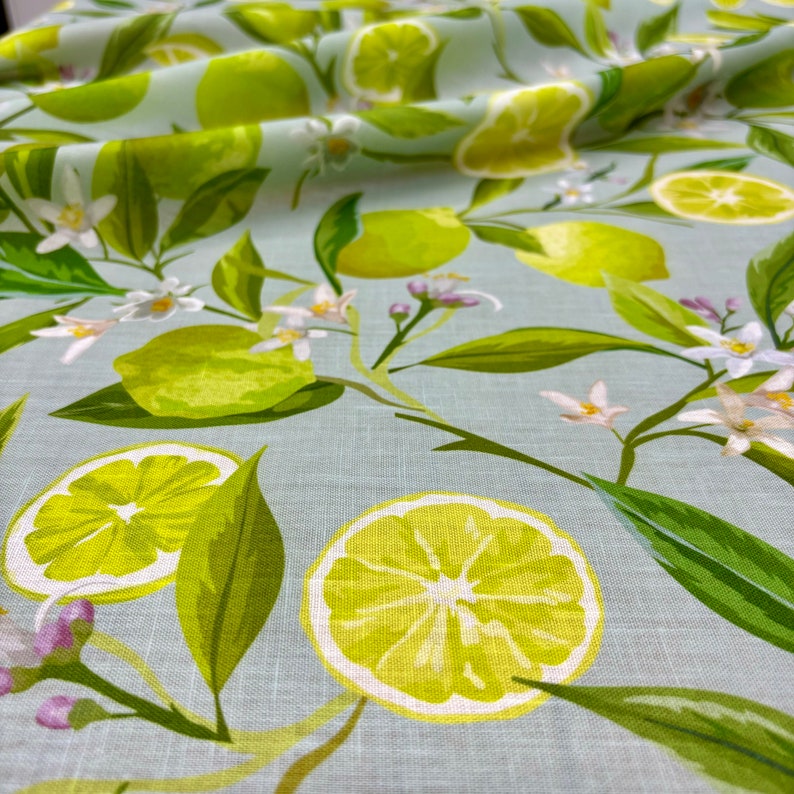 May include: Close-up of a fabric with a light blue background, featuring a repeating pattern of green limes, lime slices, white flowers, and green leaves. The fabric has a linen-like texture.