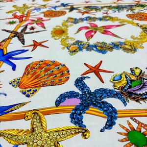 May include: White fabric with a repeating pattern of starfish, seashells, and floral elements in shades of orange, blue, gold, and pink. The design includes ornate gold borders and jewel-like accents.