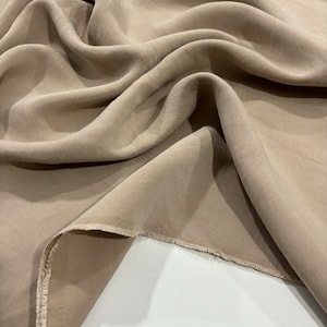 Puede incluir: Primer plano de una tela marrón claro con una textura suave y sedosa. La tela está doblada y drapeada, mostrando su caída y flujo.