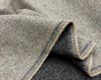 Puede incluir: Primer plano de una sección doblada de tela de lana gris. La tela tiene una superficie texturizada con un tejido visible. El borde de la tela está acabado con un ribete beige contrastante, creando un borde definido. La paleta de colores general es apagada, con tonos grises y beige.