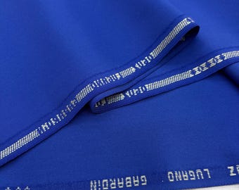 Könnte beinhalten: Ein Ballen leuchtend blauer Stoff mit silberner Bordüre. Der Stoff ist gefaltet und zeigt seine Textur und seinen Glanz. Der Text "LUGANO GABARDIN" ist am Rand des Stoffes aufgedruckt.