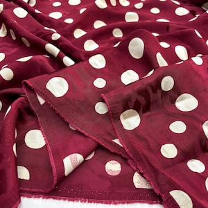 May include: Deep burgundy fabric with a repeating pattern of large, off-white polka dots. The fabric is folded, showing its texture and sheen. The material appears to be silk or satin.