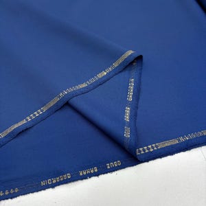 May include: A folded length of deep blue fabric with a textured finish. The edges are trimmed with a gold-coloured border. The text "OGUZ BAHAR GABARDIN" is printed along the border.