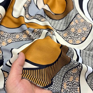 May include: A close-up shot of patterned silk fabric. The design incorporates gold, black, white, and tan in an abstract pattern. The fabric is held up, revealing its smooth texture and intricate details. The fabric is likely for clothing.