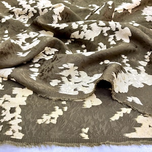 May include: Olive green fabric with a white floral design. The fabric has a textured surface and a subtle sheen. The white floral pattern looks painted or brushed on. The fabric is folded, revealing its texture and pattern.
