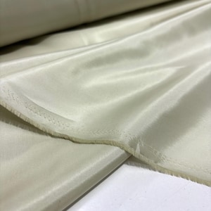May include: Close-up of a roll of pale green fabric with a silky sheen. The fabric is folded and draped, revealing a raw edge with visible stitching. The material appears soft and lightweight, suitable for apparel or home decor.