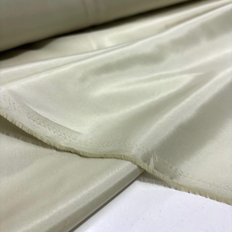 May include: Close-up of a roll of pale green fabric with a silky sheen. The fabric is folded and draped, revealing a raw edge with visible stitching. The material appears soft and lightweight, suitable for apparel or home decor.
