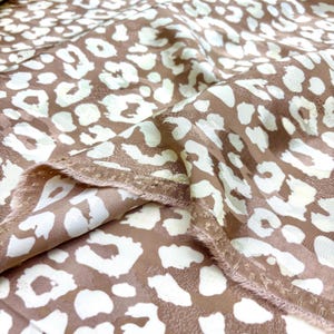 May include: Close-up of a fabric with a leopard print pattern. The fabric is a light brown colour with white and off-white spots. The edges of the fabric are frayed.