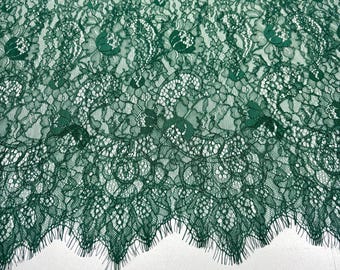 May include: Close-up of emerald green lace fabric. The delicate textile features an intricate floral pattern with a scalloped edge. The lace is semi-sheer, revealing the background. Ideal for sewing projects, apparel, or home decor.