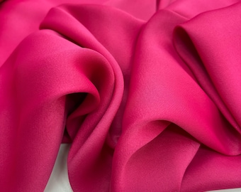 Puede incluir: Primer plano de una tela de color rosa vibrante con una textura suave y sedosa. La tela está drapeada y doblada, mostrando su calidad fluida y lujosa.