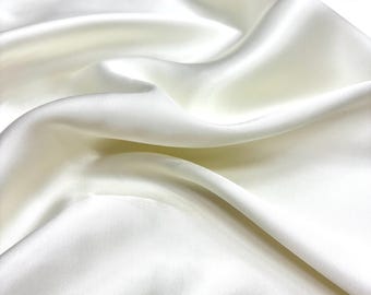 Puede incluir: Tela de satén color marfil con pliegues suaves y un brillo lustroso. La superficie de la tela refleja la luz, creando reflejos y sombras. El material parece suave y fluido, adecuado para proyectos de ropa o decoración del hogar.