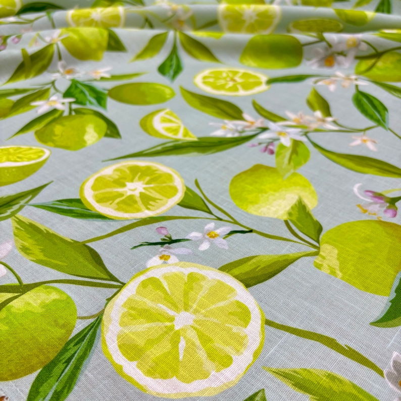 May include: Fabric with a light blue background featuring a pattern of green limes, lime slices, green leaves, and white flowers. The limes are in various sizes and orientations, creating a fresh, summery design.