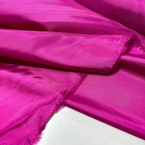 May include: Close-up of vibrant fuchsia fabric. The material is smooth and appears to be silk or satin, with visible folds and a slight sheen. The edges of the fabric are unfinished, showing the raw texture.