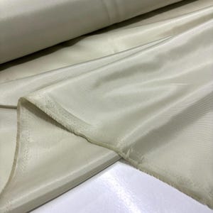May include: A roll of pale yellow fabric with a silky sheen. The fabric is draped, showing folds and a raw edge. The material appears to be lightweight and suitable for clothing or decorative projects.