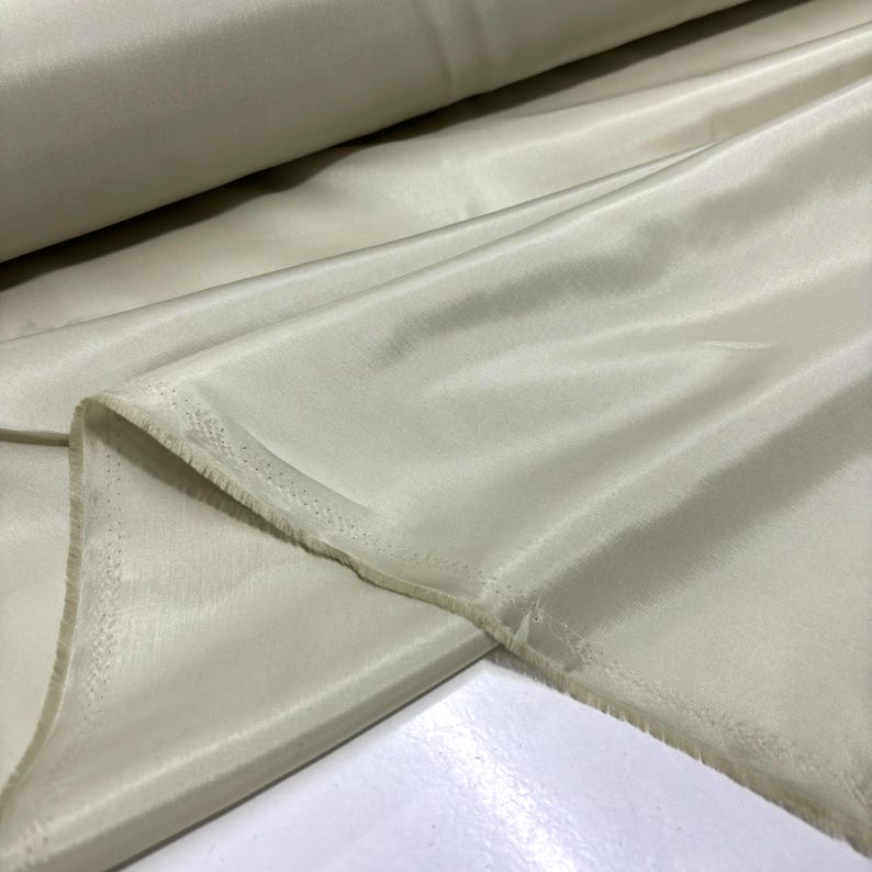 May include: A roll of pale yellow fabric with a silky sheen. The fabric is draped, showing folds and a raw edge. The material appears to be lightweight and suitable for clothing or decorative projects.