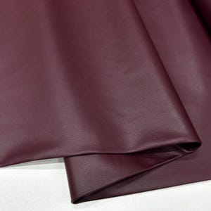 Puede incluir: Primer plano de una tela de imitación de cuero de color burdeos. La tela tiene una superficie texturizada y está doblada por la mitad.