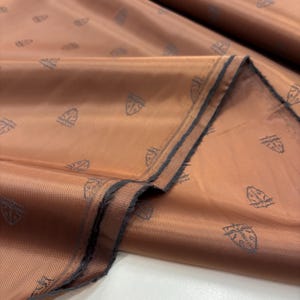 Puede incluir: Primer plano de una tela doblada en un cálido tono cobre. El material tiene un brillo sutil y está estampado con pequeños motivos repetidos en forma de escudo en un gris más oscuro. Los bordes de la tela son visibles, mostrando un borde más oscuro.