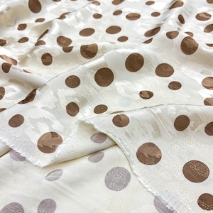 May include: Cream-coloured fabric featuring a repeating pattern of brown polka dots in various sizes. The fabric has a subtle floral design in the background. The material is folded, showing the front and back.