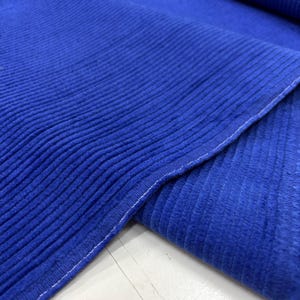 Puede incluir: Primer plano de una vibrante tela de pana azul. La tela tiene una textura acanalada con líneas verticales visibles. Los bordes están cuidadosamente acabados con costuras blancas, y la tela está doblada sobre sí misma.