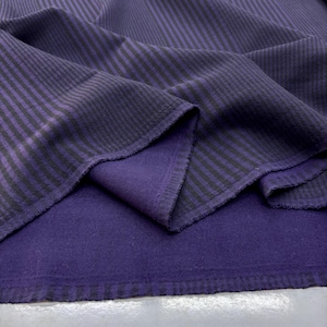 Peut inclure: Gros plan sur un tissu plié avec un motif rayé. La couche supérieure est violet foncé avec des rayures verticales noires. Le dessous révèle un tissu violet foncé uni. Les bords du tissu sont visibles, montrant les couches.