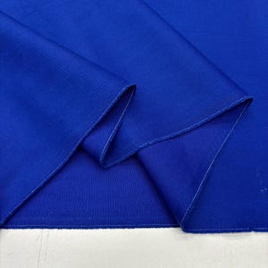 Puede incluir: Primer plano de una tela azul vibrante con textura visible y costuras blancas en los bordes. La tela está doblada, creando sombras y resaltando la profundidad del material. La imagen muestra el color y el tejido textil.