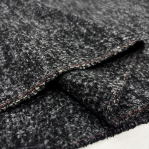 Può includere: Primo piano di un tessuto strutturato nelle tonalità del grigio e del nero. Il materiale ha un aspetto maculato e una cucitura visibile con cuciture rosse e verdi. Il tessuto è piegato, rivelando la sua costruzione a strati.