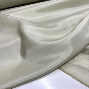 May include: A close-up of a fabric with a silky, lustrous appearance. The fabric is a pale, creamy color, with soft folds and subtle variations in tone. The material appears smooth and flowing, suggesting it is lightweight and drapes well.