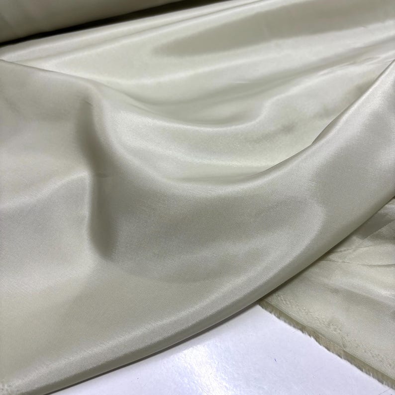 May include: A close-up of a fabric with a silky, lustrous appearance. The fabric is a pale, creamy color, with soft folds and subtle variations in tone. The material appears smooth and flowing, suggesting it is lightweight and drapes well.