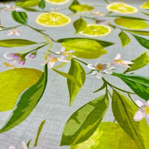 May include: Close-up of a fabric with a light blue background featuring a pattern of green limes, lime slices, white blossoms, and green leaves. The design is a repeating pattern of citrus fruit and floral elements.