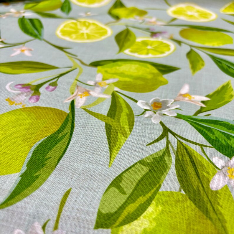 May include: Close-up of a fabric with a light blue background featuring a pattern of green limes, lime slices, white blossoms, and green leaves. The design is a repeating pattern of citrus fruit and floral elements.