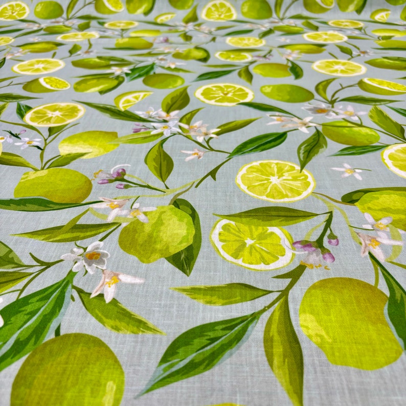 May include: Fabric with a repeating pattern of green limes, lime slices, white blossoms, and green leaves on a light blue background. The design features whole limes, sliced limes, and floral elements.