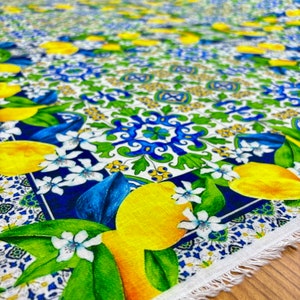 May include: White fabric with a blue, green, and yellow pattern. The pattern features lemons, leaves, and flowers. The fabric has a fringed edge.
